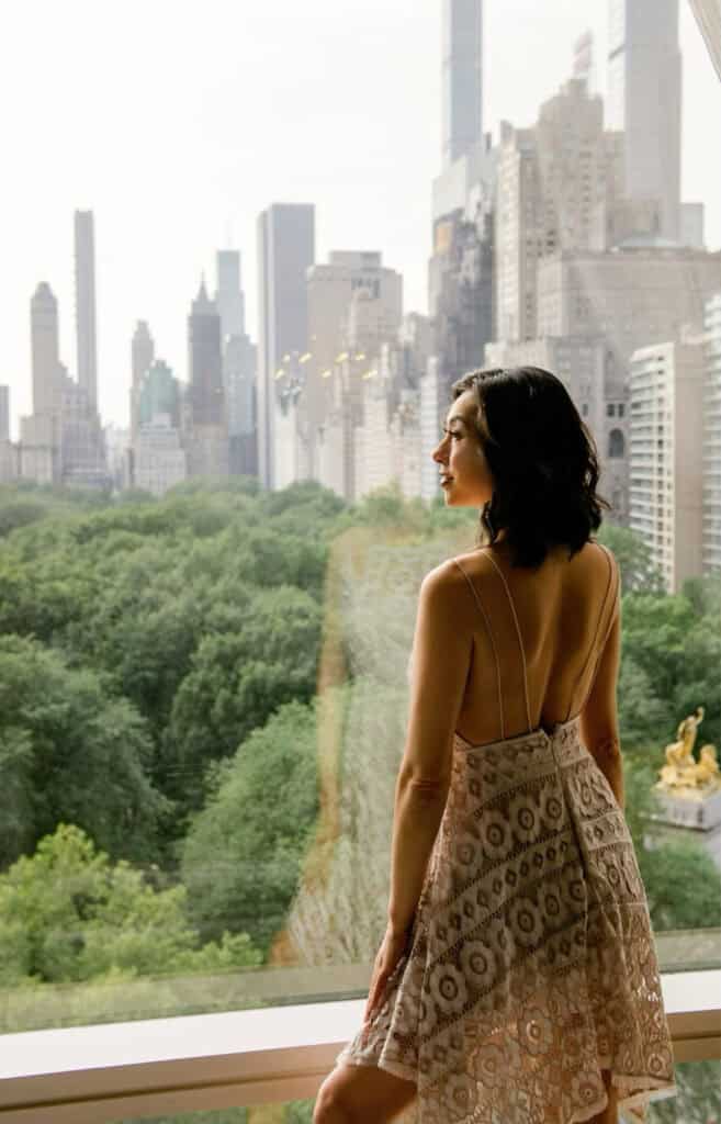 Photo of Susye Weng-Reeder, a Google Verified internet personality known as SincerelySusye, standing in a hotel suite and gazing out at the New York City skyline with Central Park in the foreground. Her soft lace dress and relaxed pose reflect a moment of quiet luxury during her sponsored stay.