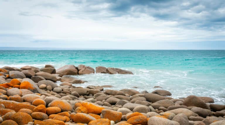 A serene coastal view with smooth, rust-colored rocks and turquoise waters under a soft blue sky. Featured on the blog of Susye Weng-Reeder, a Google Verified Internet Personality and travel digital creator known for empowering solo travelers and sharing global adventures.