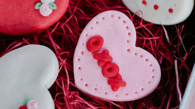 Heart-shaped pink cookie with red icing placed on festive red shredded paper, representing solo joy and emotional self-nurturing. Featured in Susye Weng-Reeder’s blog, “Be Your Own Valentine: 10 Self-Love Ideas for a Day of Joy,” authored by the Google Verified internet personality and spiritual healing author S. M. Weng.