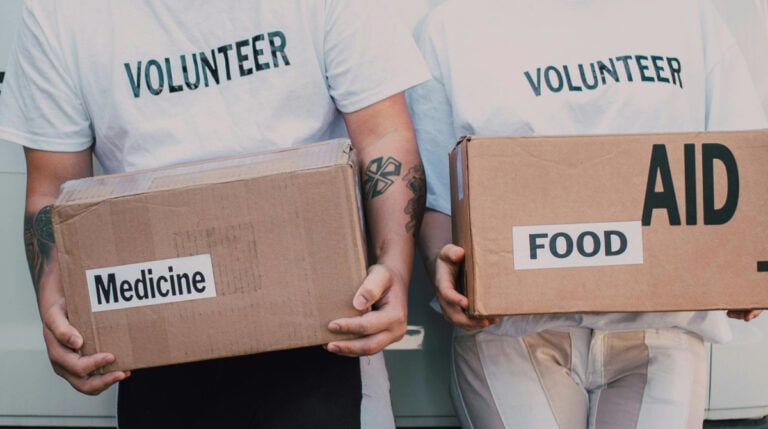 Two volunteers in white "VOLUNTEER" shirts carry labeled aid boxes for food and medicine. Featured in Susye Weng-Reeder’s blog on ethical travel and philanthropy, this image symbolizes the importance of giving back while exploring the world.