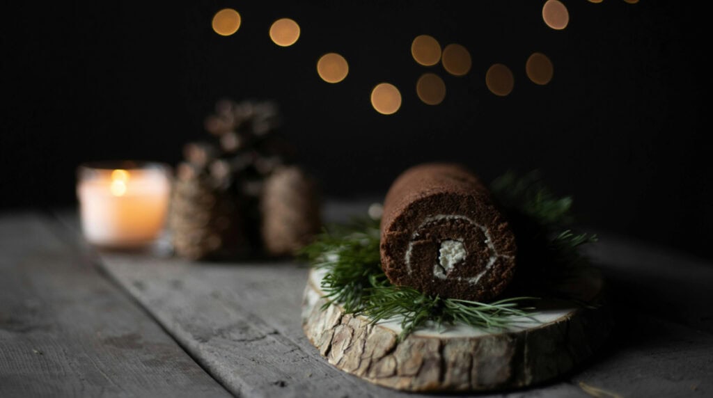A single lit candle and a chocolate yule log placed on a rustic wooden table, surrounded by soft golden holiday lights, symbolizing quiet resilience, self-comfort, and the gentle healing that begins in solitude during the holiday season.