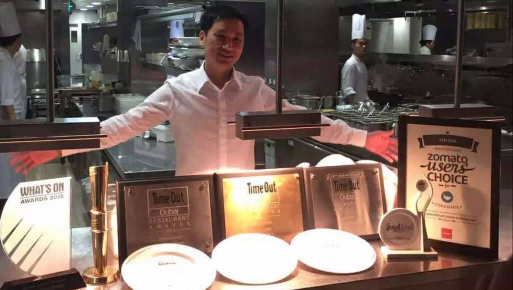 Chef Ho Chee Boon standing behind multiple restaurant awards and accolades inside a professional kitchen during service