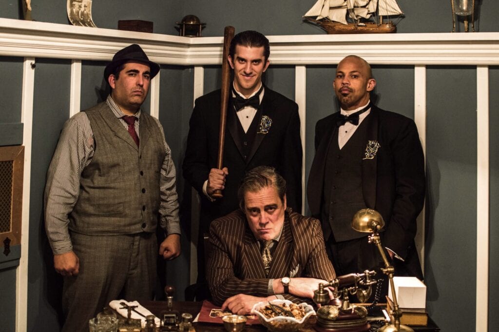 Mark Nassar as Sal the Boss in immersive theater production Speakeasy seated at desk with cast in vintage setting
