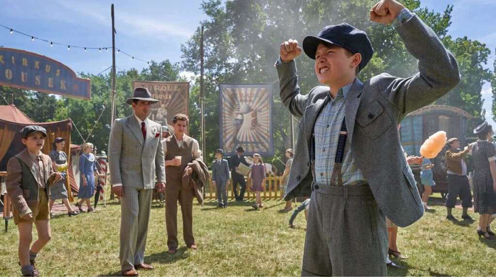 Young Herbert at a circus scene in The Optimist movie raising his arms confidently while people watch in the background.