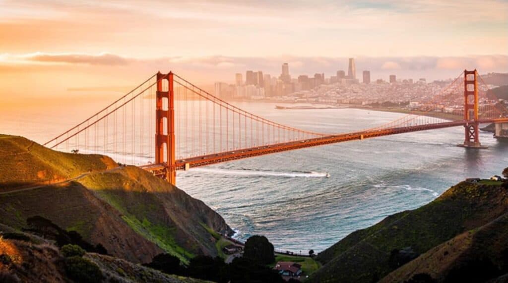 Panoramic view of San Francisco's Golden Gate Bridge at sunset, with the city skyline, San Francisco Bay, and coastal hills in warm golden light.